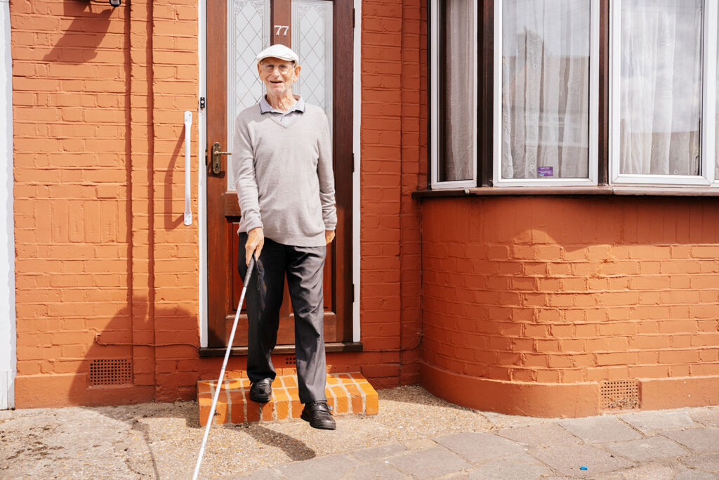 A picture of an MAB client, Parry. Standing outside his front door, with a white cane and a lovely smile on his face.